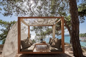 Un gazebo in legno con un tavolo e sedute imbottite, affacciato su uno specchio d'acqua e un paesaggio costiero.