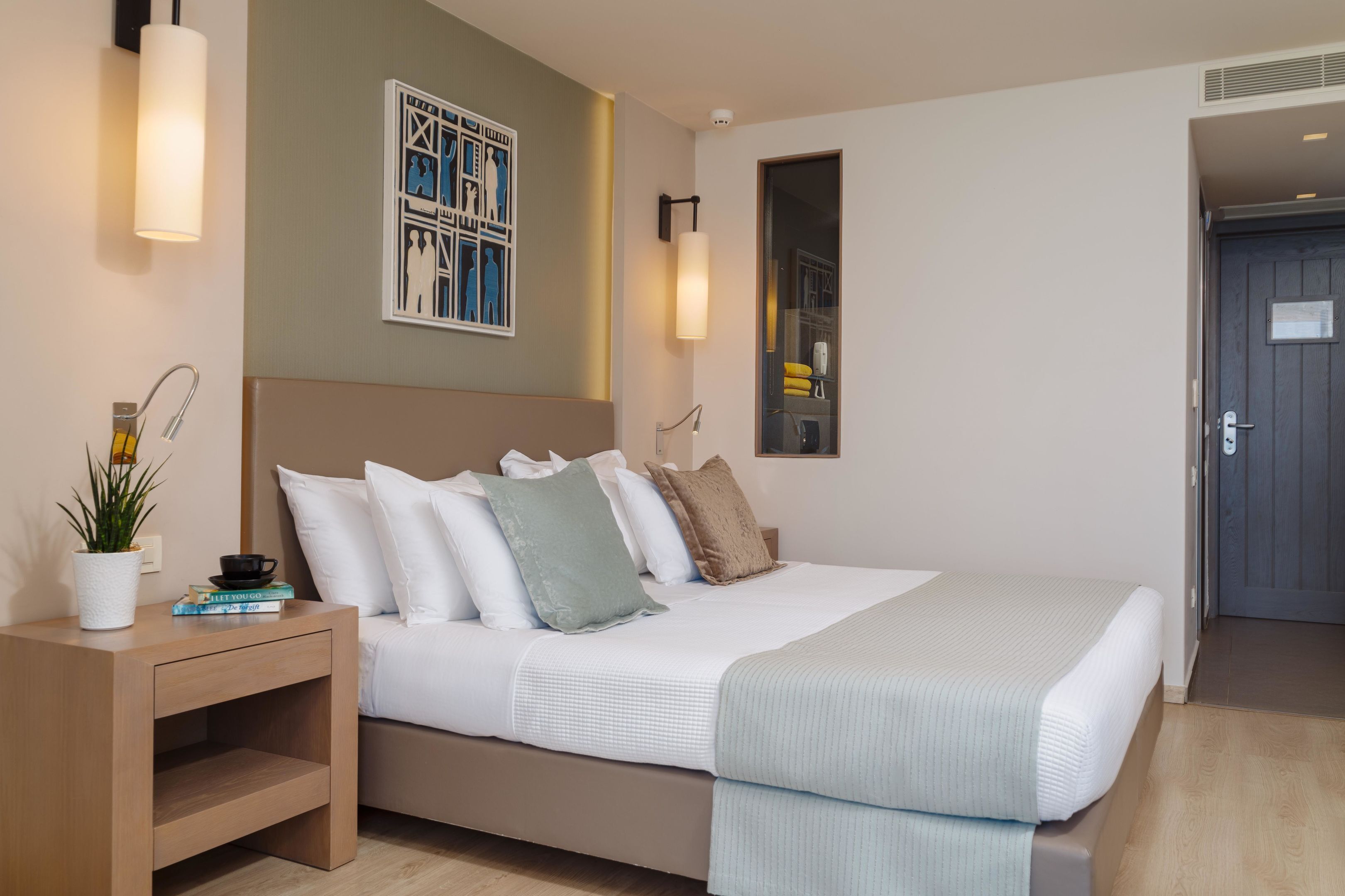 A modern hotel room with a large bed, a bedside table with a plant, a wall-mounted artwork, and a door with a sign.