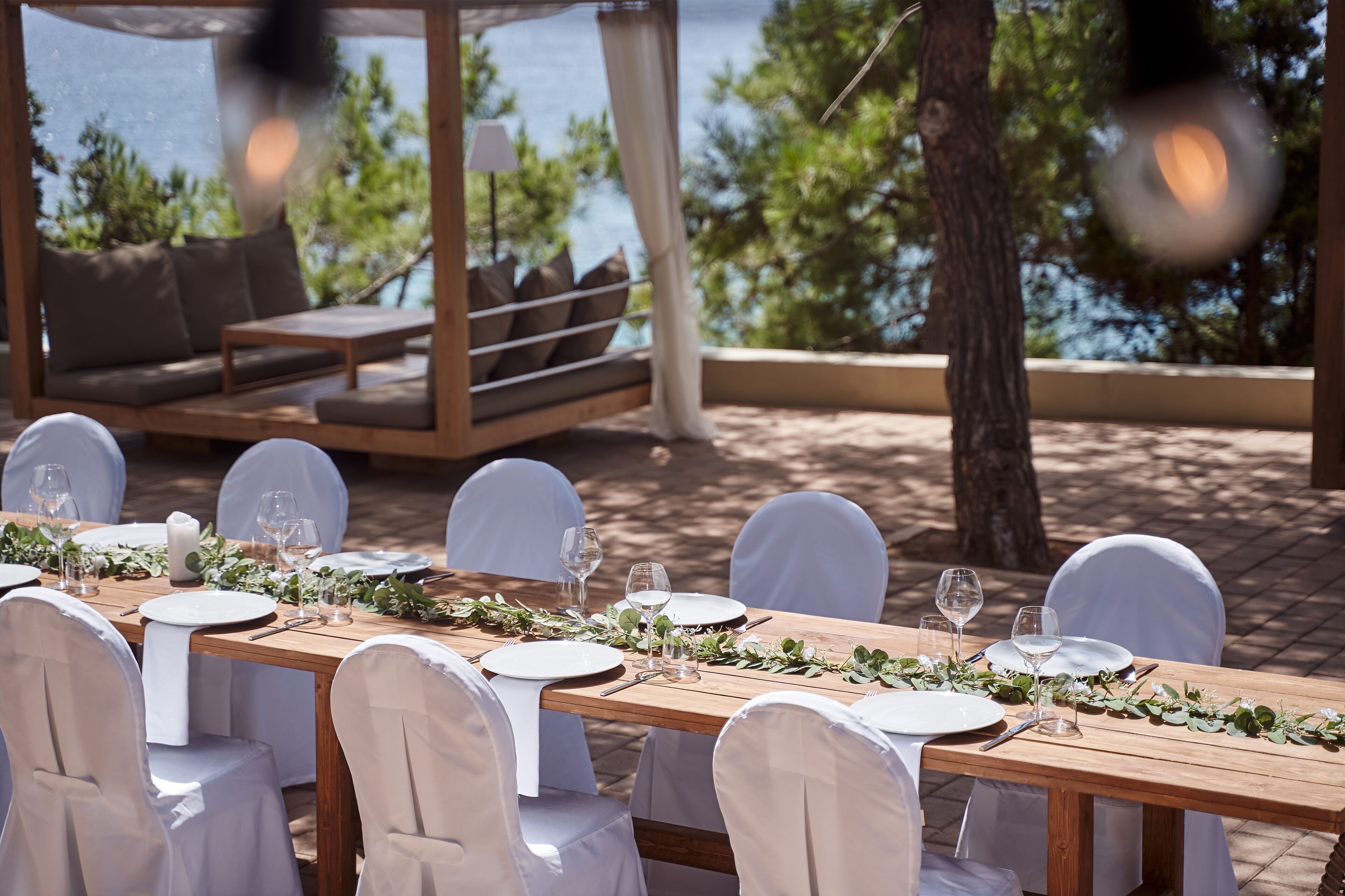 A wooden dining table set with white chairs, plates, and wine glasses, surrounded by greenery and overlooking a body of water.