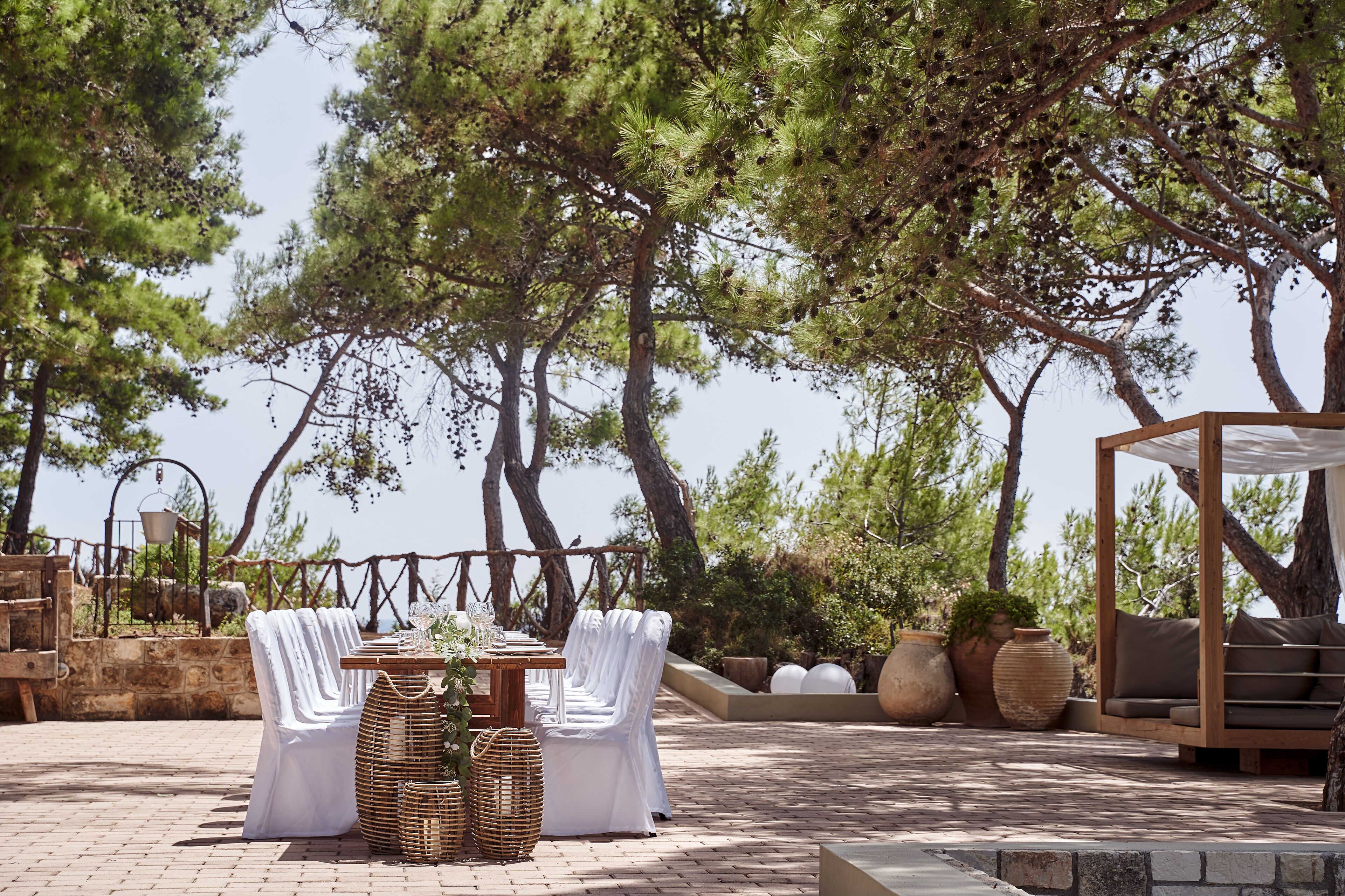 An outdoor dining area with a table set for two, surrounded by white-covered chairs, a wooden pergola, and potted plants.