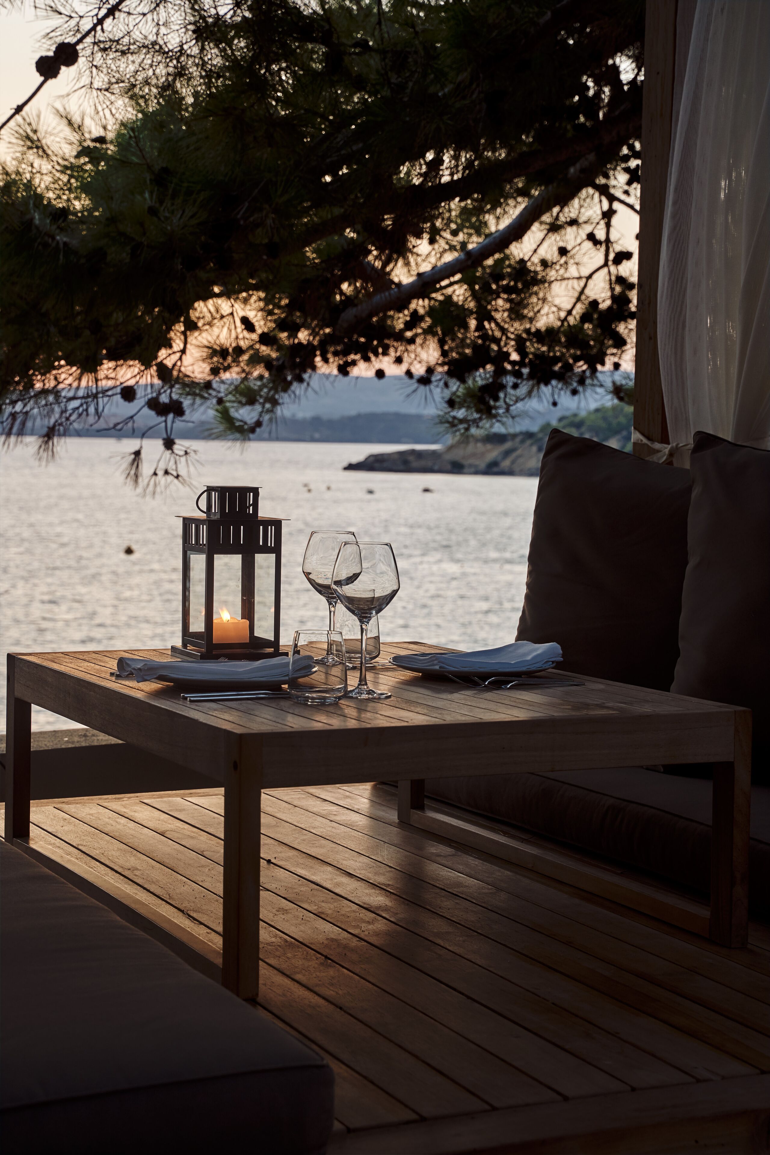 Una mesa de madera con una vela, copas de vino y un farol en una terraza con vista a un cuerpo de agua.
