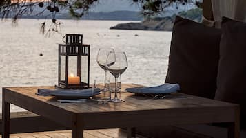 Una mesa de madera con una vela, copas de vino y un farol en una terraza con vista a un cuerpo de agua.