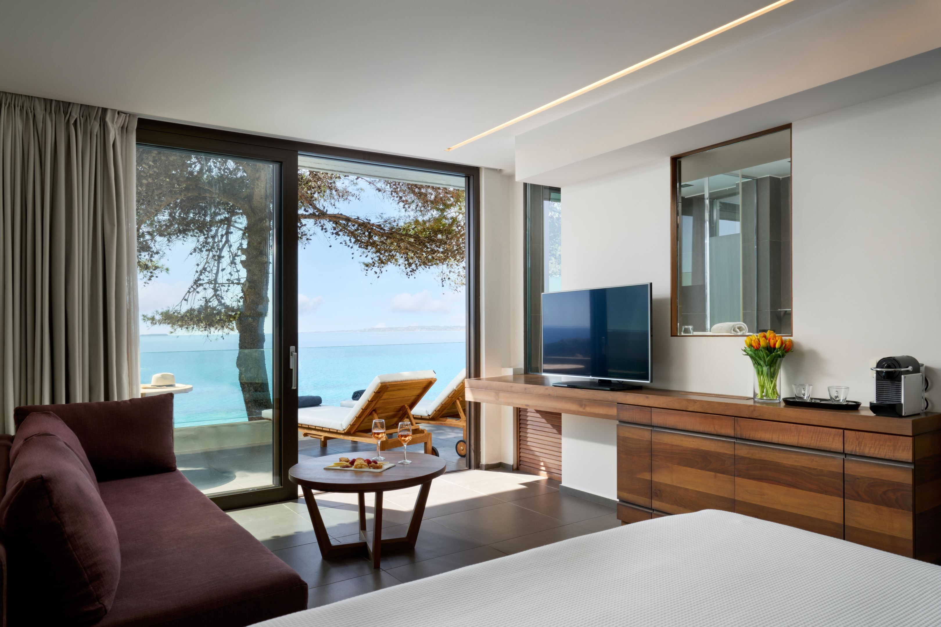 A modern hotel room with a large bed, a sofa, a desk with a TV, and a view of the sea.