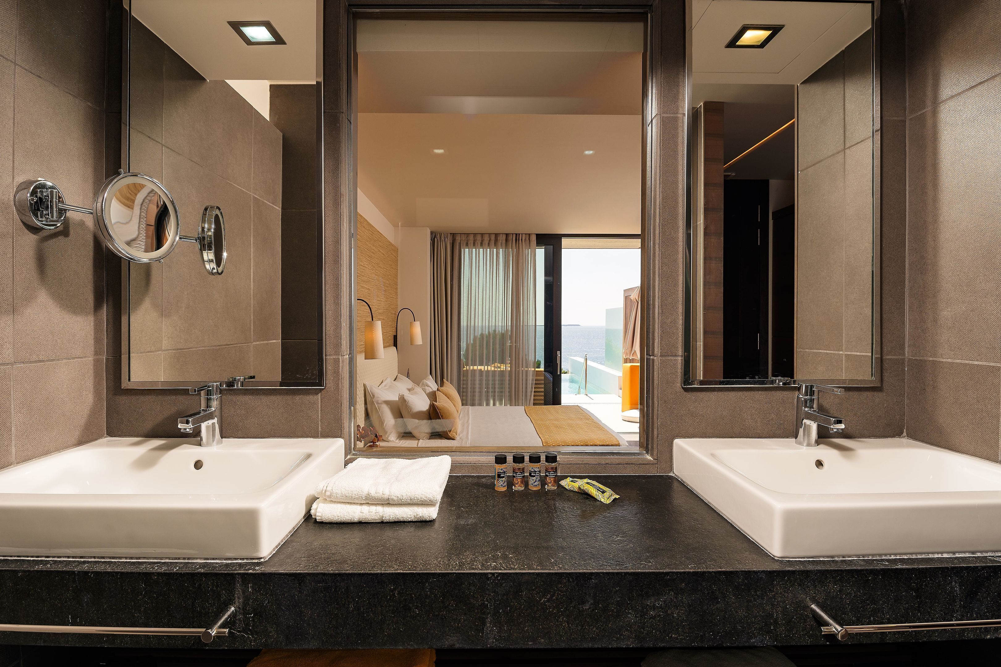 A modern bathroom with two sinks, a large mirror, and a view of a bedroom with a balcony.