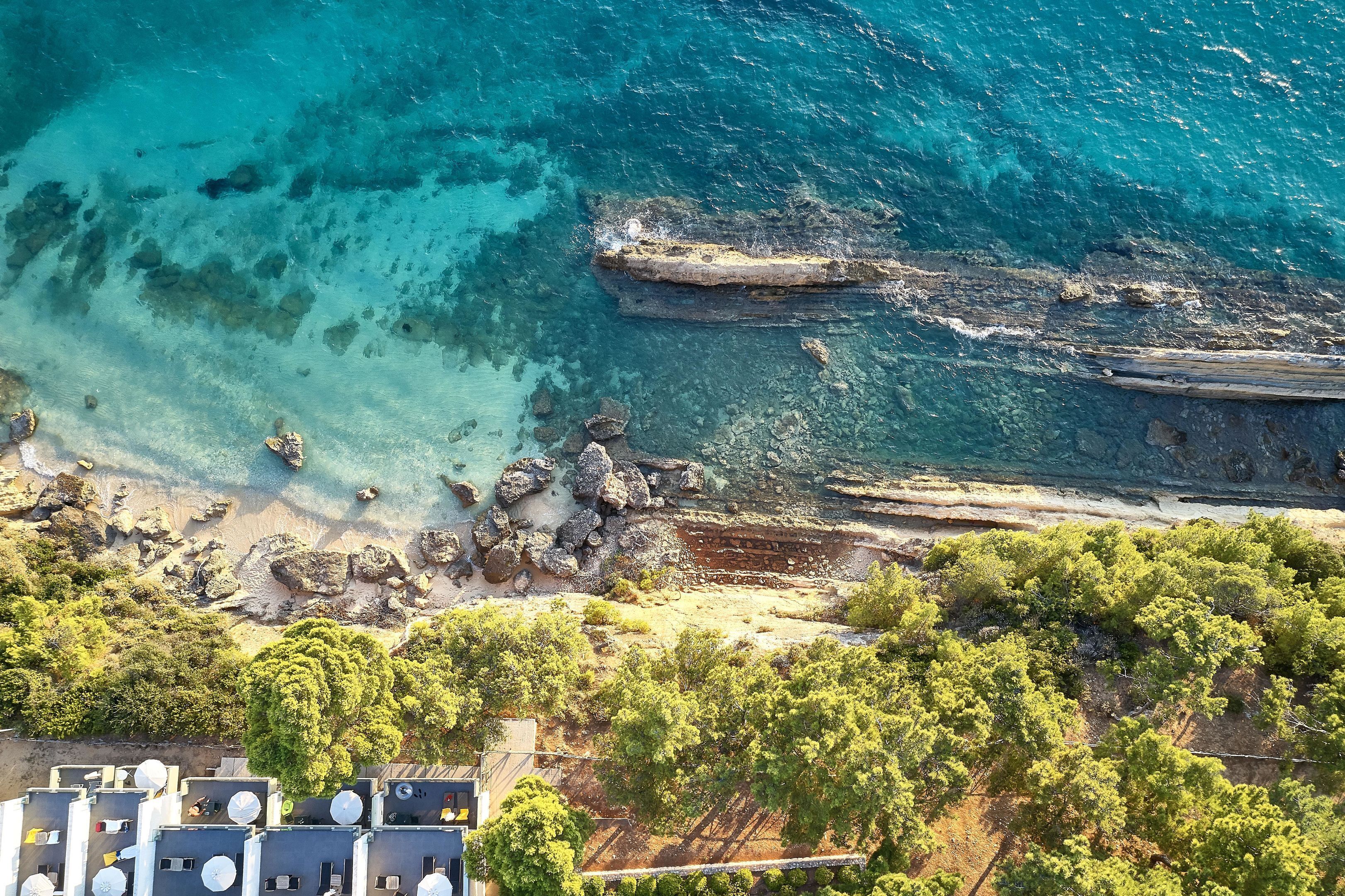 An aerial view of a coastal area with rocky terrain, clear turquoise waters, and dense green vegetation.