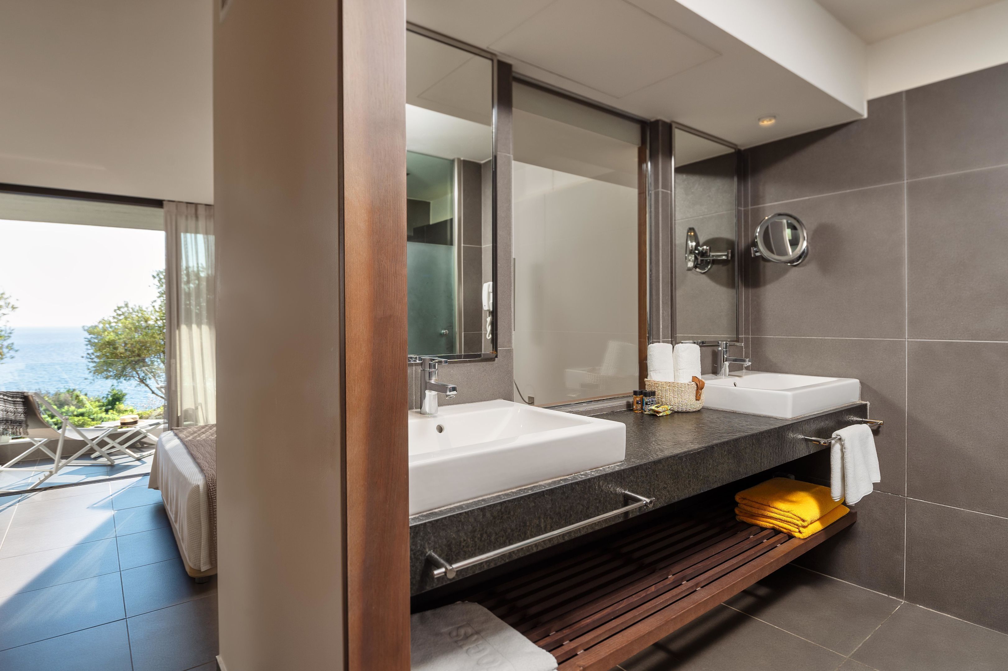 A modern bathroom with two sinks, a large mirror, and a view of the sea.