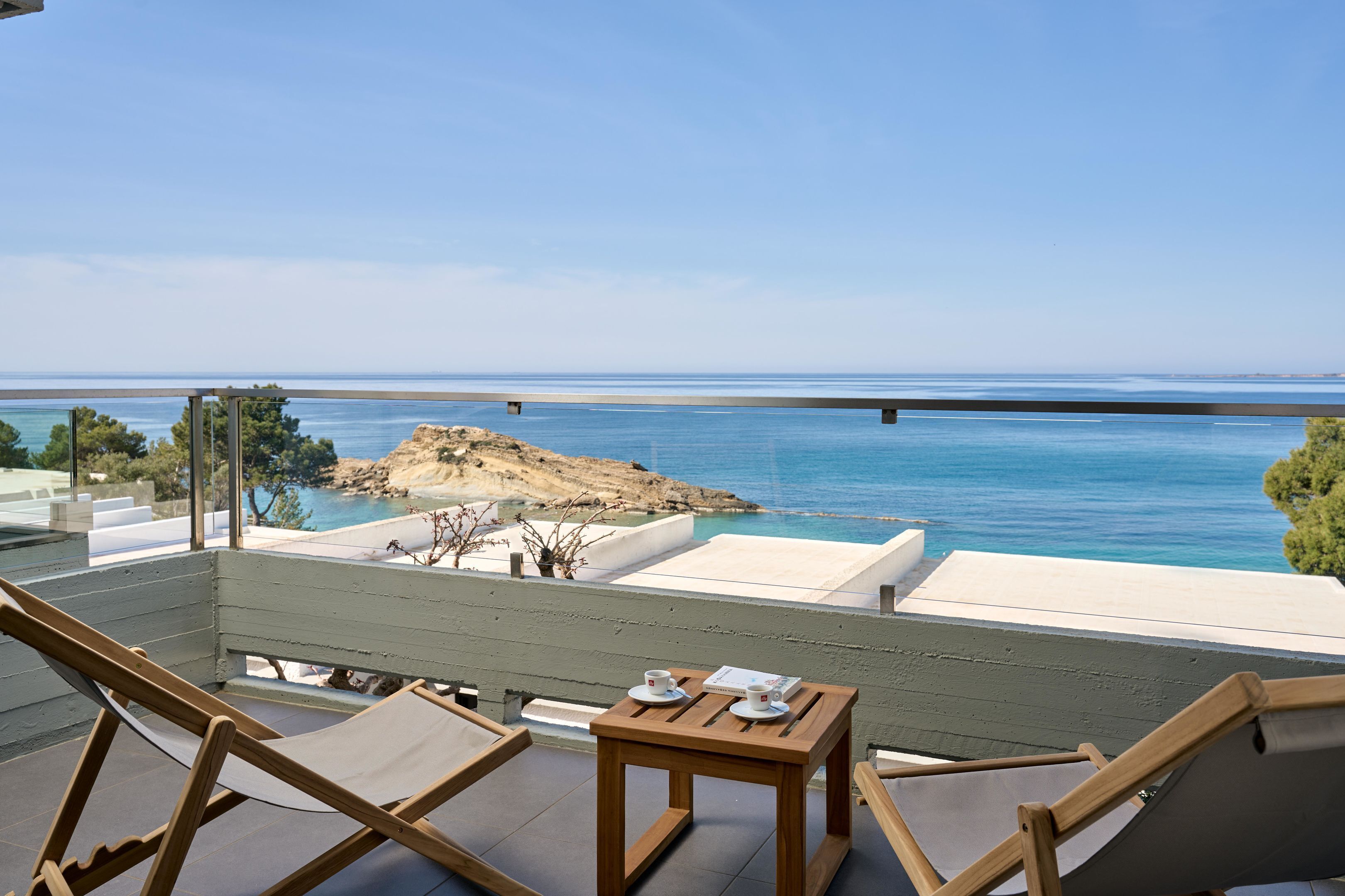 Un balcon avec vue sur la mer, des chaises en bois, une petite table avec une tasse et une soucoupe, et un espace détente blanc.