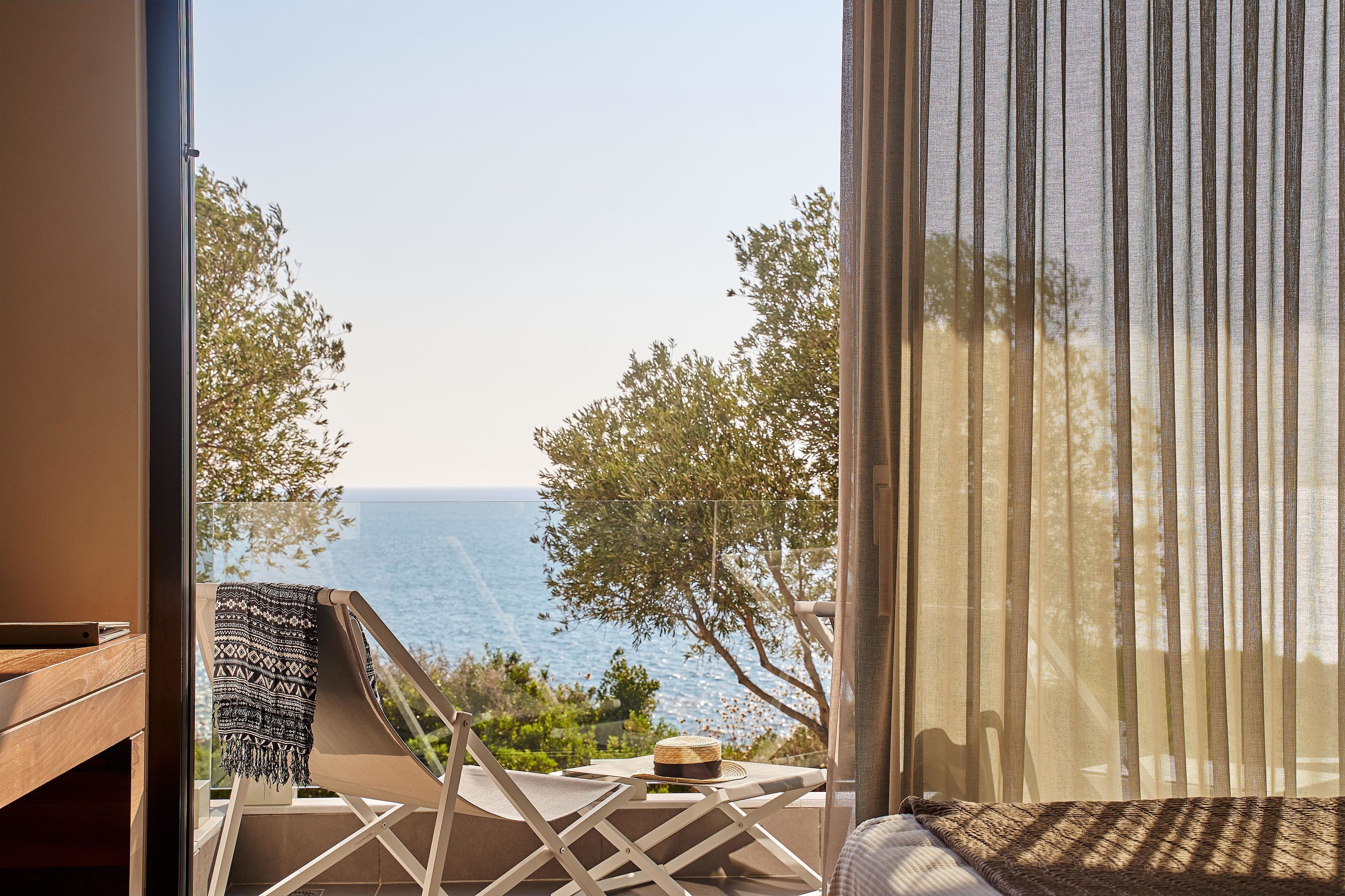 A balcony with a view of the sea, a chair, a table, and a window with sheer curtains.