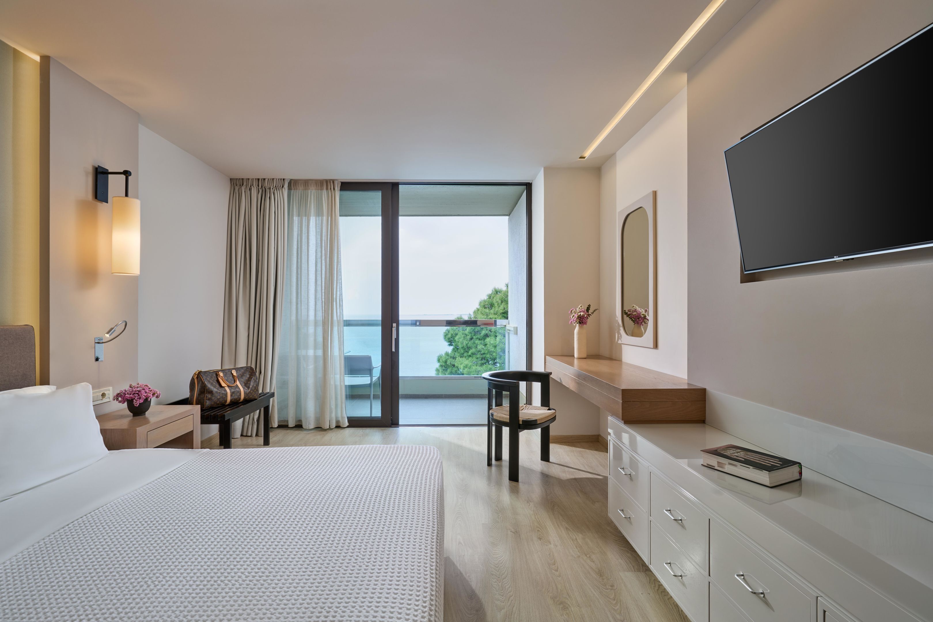 Quarto de hotel moderno com uma cama grande, uma secretária com uma cadeira, uma TV de tela plana, uma janela ampla com cortinas e uma varanda com vista.