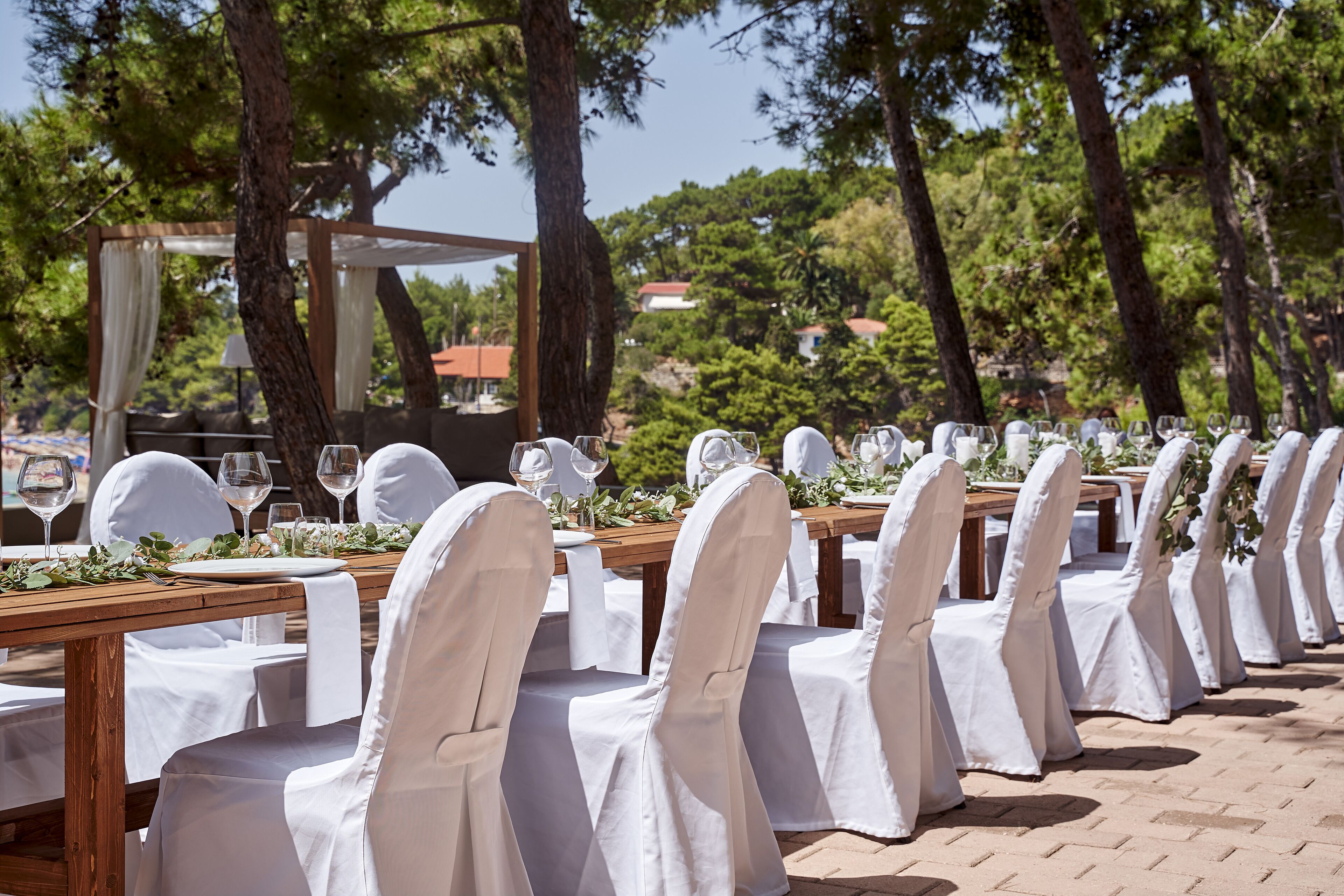 Un área de comedor al aire libre con sillas y mesas cubiertas de blanco, con árboles y cielo despejado de fondo.