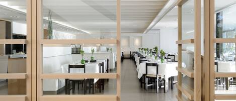Un comedor moderno con mesas y sillas blancas, separadores de madera y plantas en macetas.