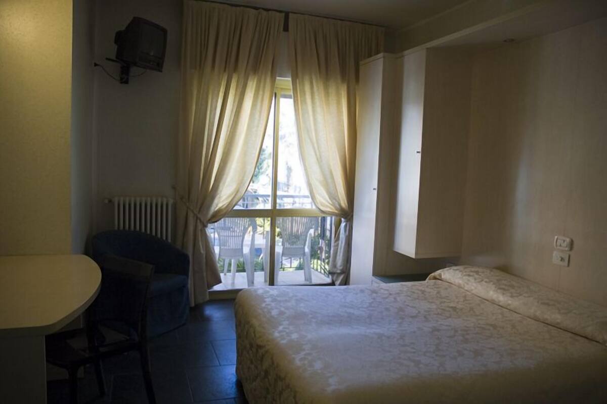 A hotel room with a bed, a small table, a chair, and a window with curtains.