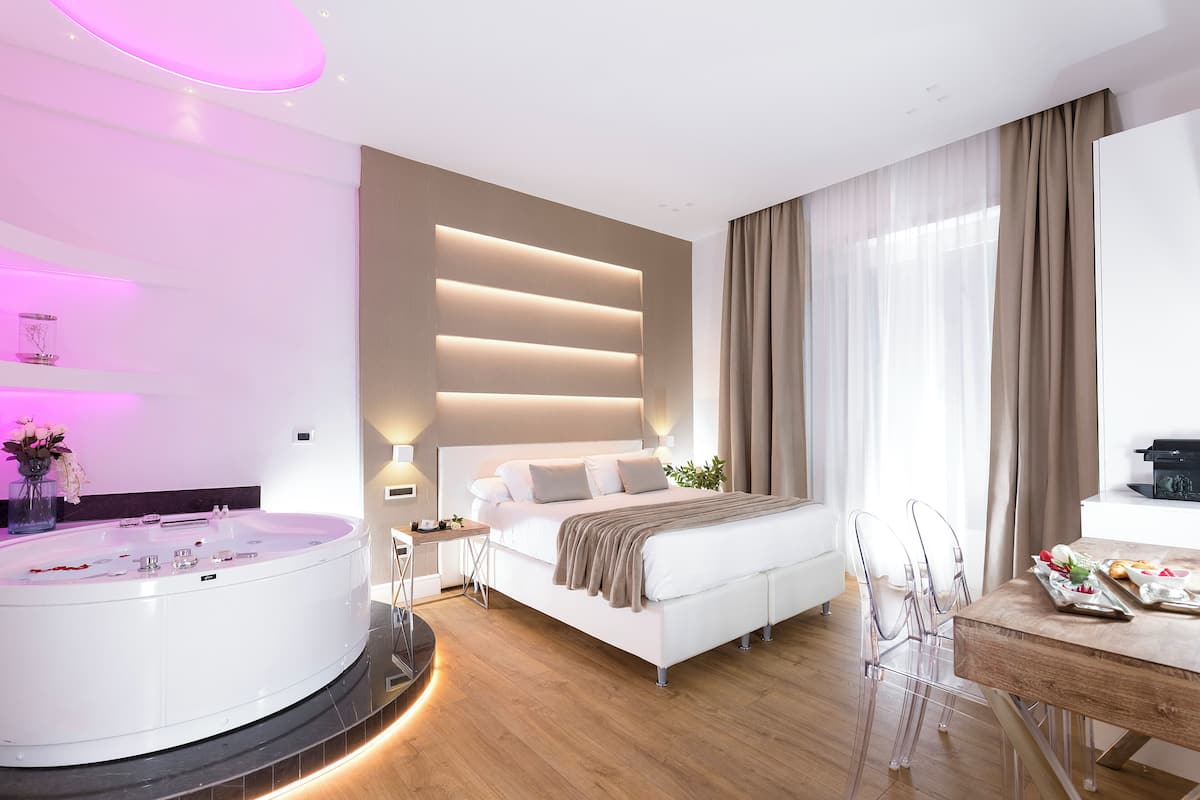 A modern bedroom with a large bed, a built-in bathtub, and a dining area with clear chairs.