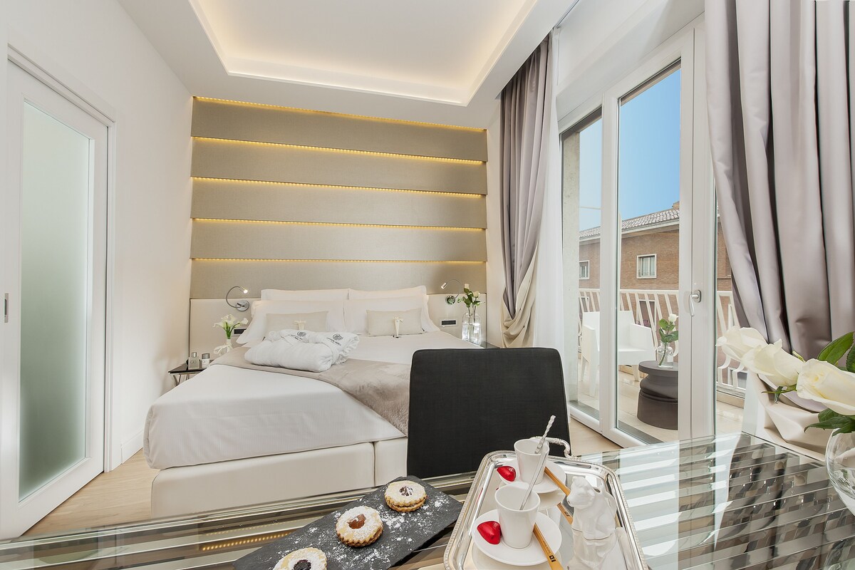 A modern hotel room with a large bed, a sitting area with a glass table, and a balcony with a view.