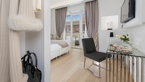 A modern hotel room with a bed, a glass table with a chair, a TV, and a view of the city through large windows.