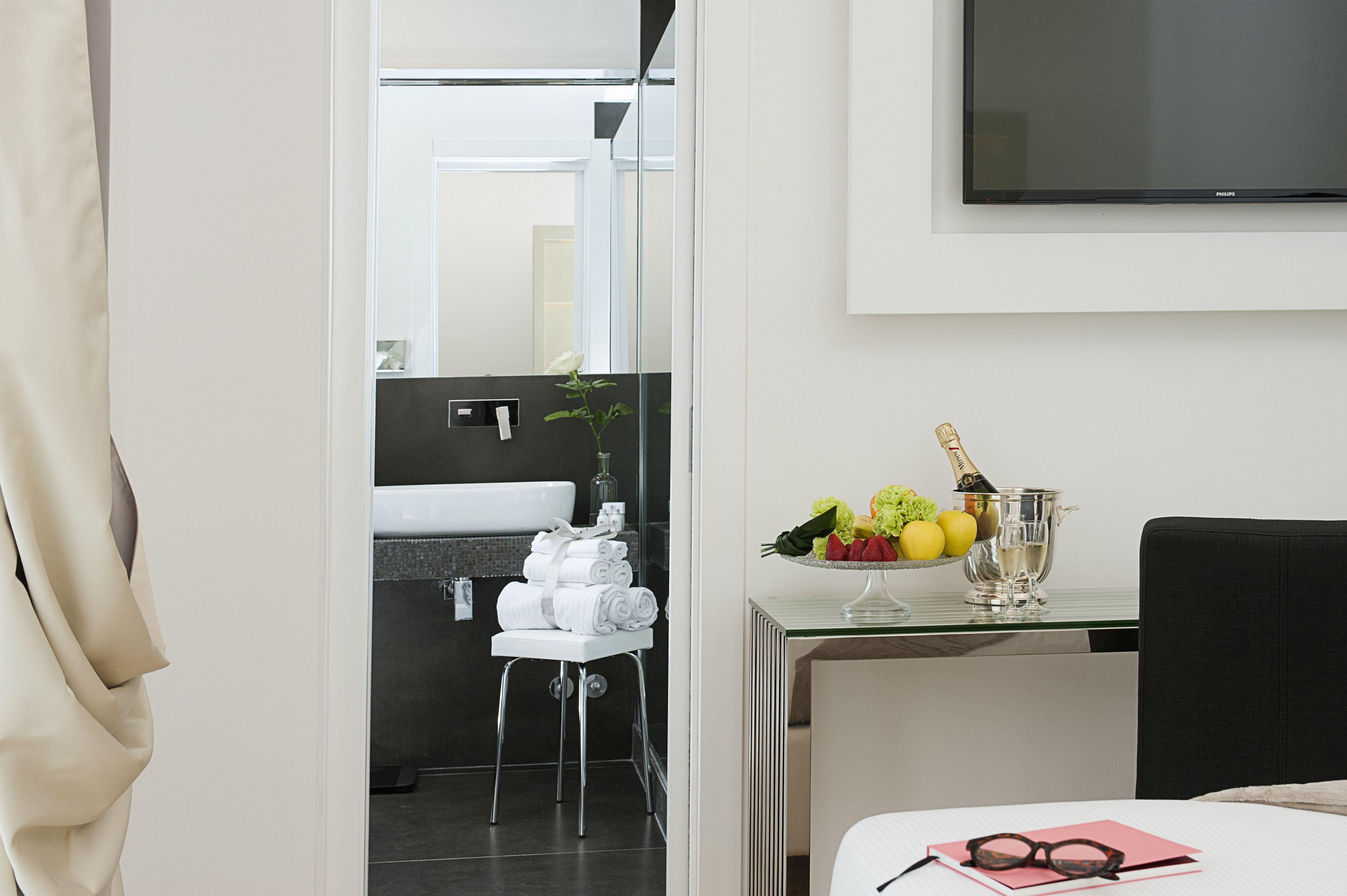A hotel room with a modern bathroom, a dining area with a table set for two, and a flat-screen TV mounted on the wall.