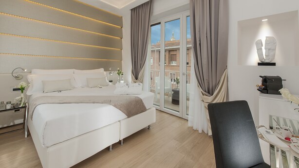 A modern hotel room with a large bed, a desk with a chair, a glass door leading to a balcony, and a sculpture on the wall. - Vatican Style (Rome)