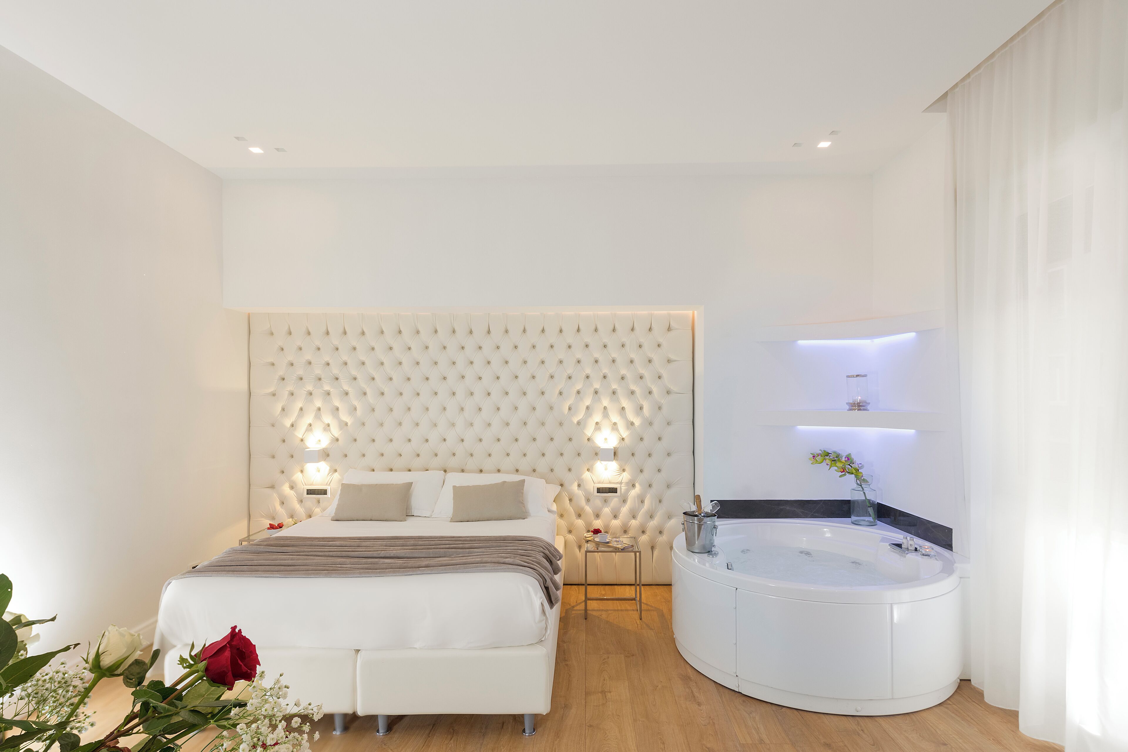 Une chambre moderne avec un grand lit, un luminaire encastré fixé au mur, une petite table de chevet et une salle de bain séparée, dotée d’une baignoire et d’un miroir.