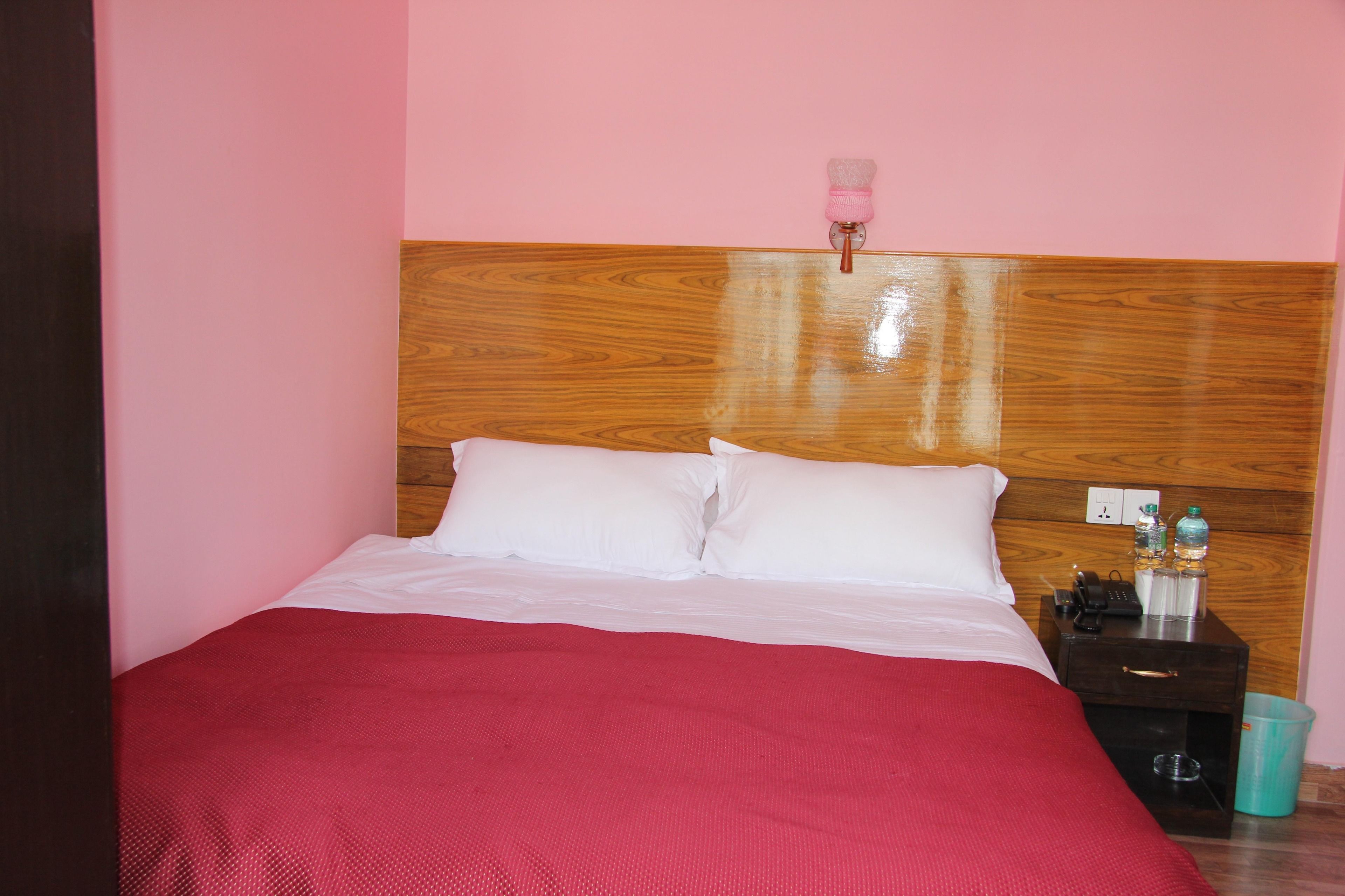 A bed with white linens and a red blanket, a wooden headboard, a bedside table with a phone, a bottle, and a trash bin.