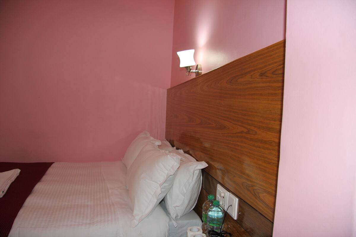 A hotel room with a wooden headboard, a bed with white linens, a nightstand with a lamp and a bottle of water, and a wall socket.