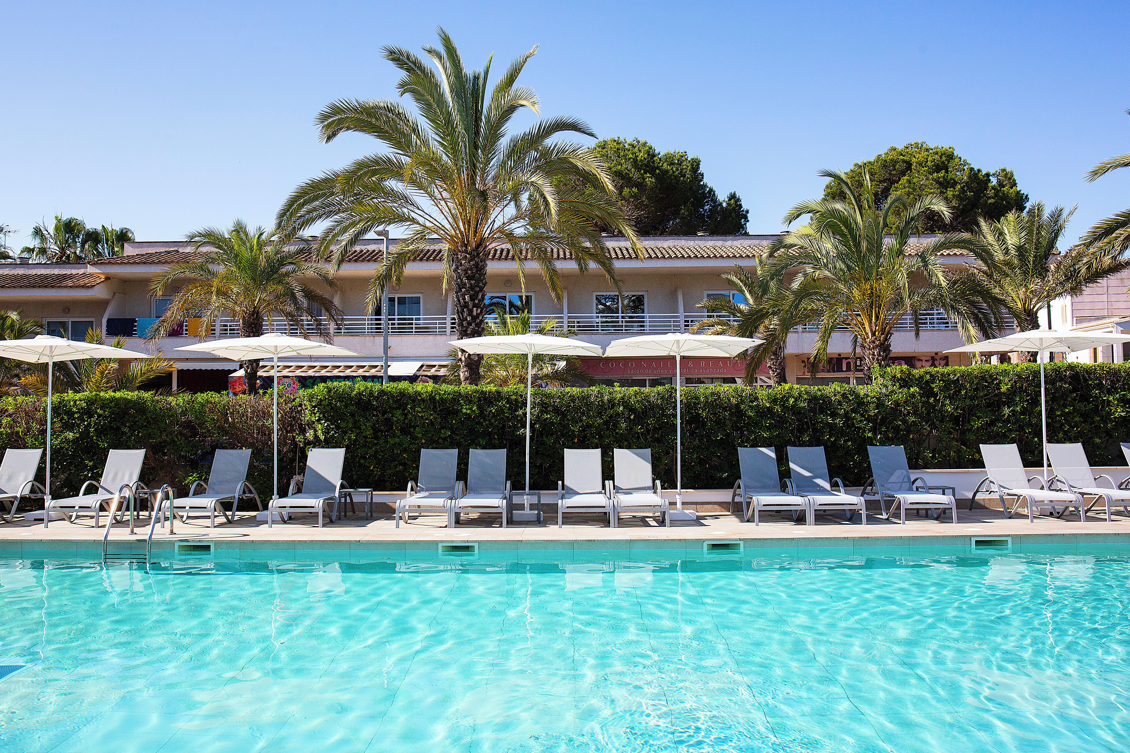 A pool area with lounge chairs and umbrellas, palm trees, and a building with a tiled roof in the background.