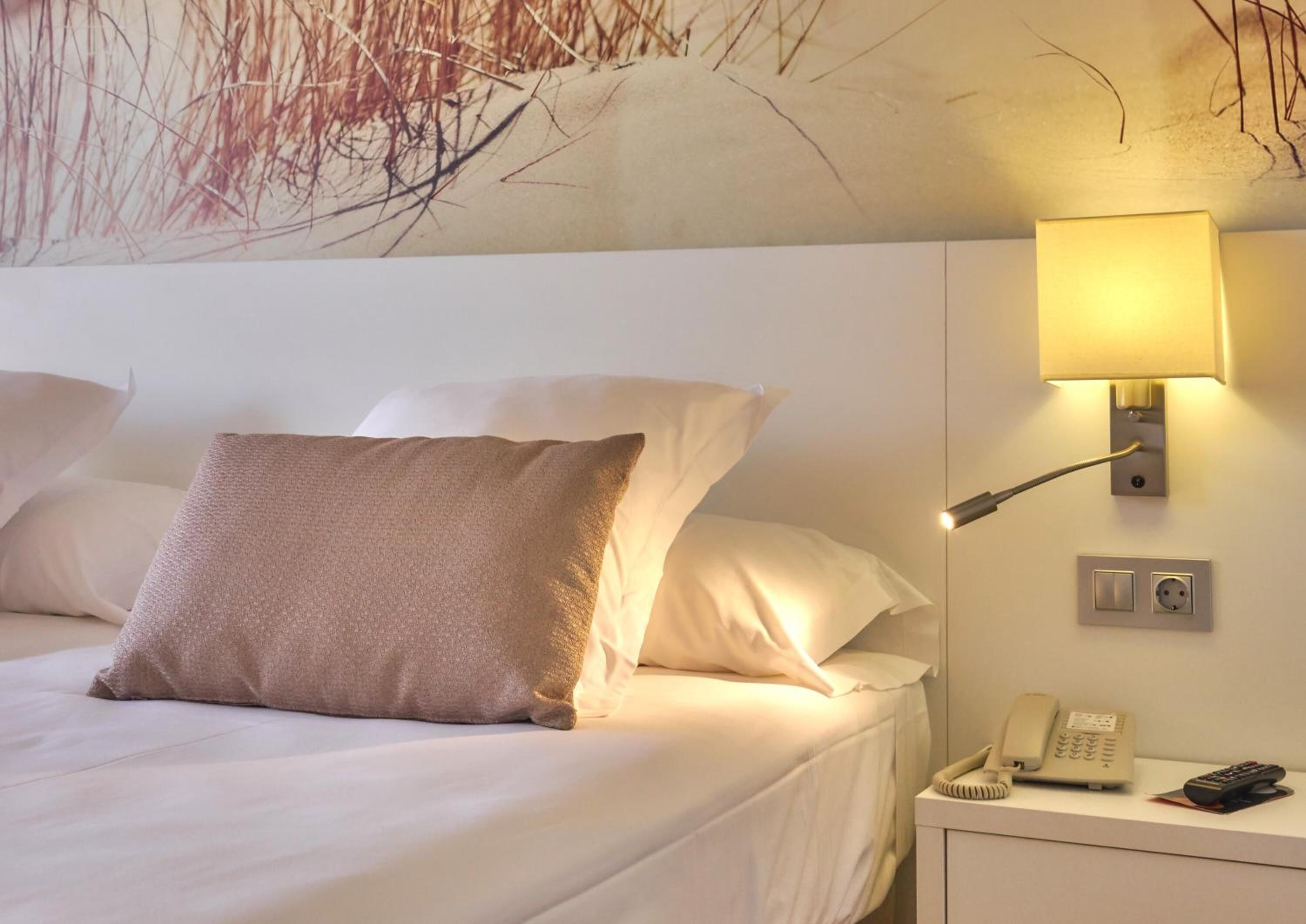 Habitación de hotel con cama, almohadas, mesita de noche con lámpara, teléfono y aplique de pared.