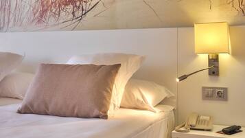 Habitación de hotel con cama, almohadas, mesita de noche con lámpara, teléfono y aplique de pared.