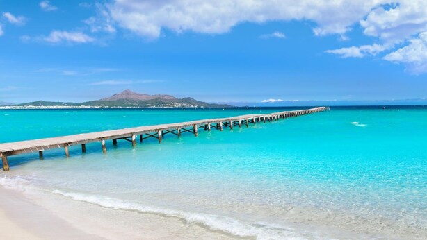 A beach with clear turquoise water, a wooden pier extending into the sea, and a distant mountain range under a partly cloudy sky. - BQ Sarah Hotel - Adults Only (Santa Margalida)