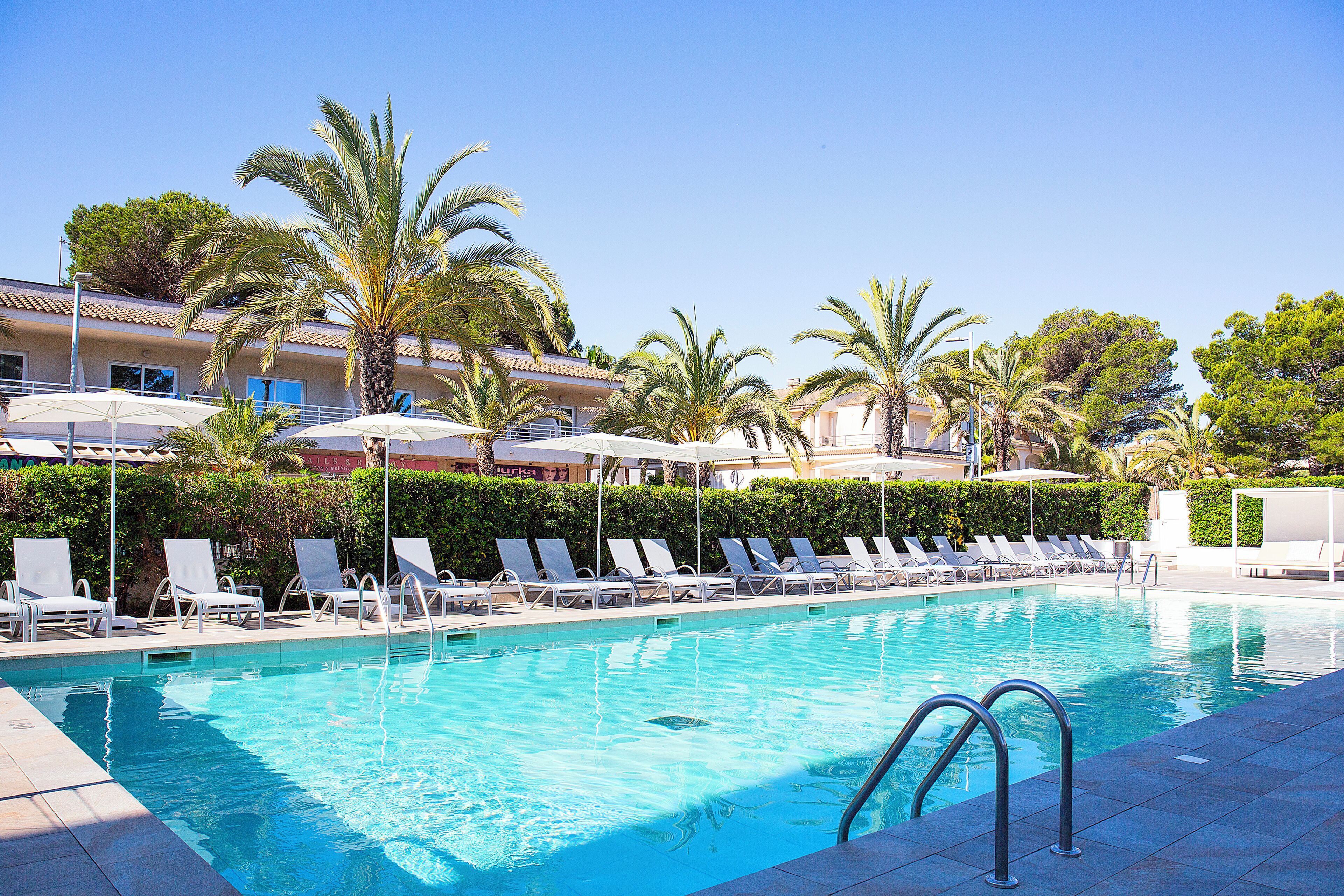 A swimming pool with lounge chairs and umbrellas, surrounded by palm trees and a building with balconies.
