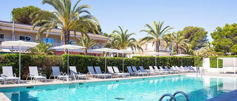 A swimming pool with lounge chairs and umbrellas, surrounded by palm trees and a building with balconies.