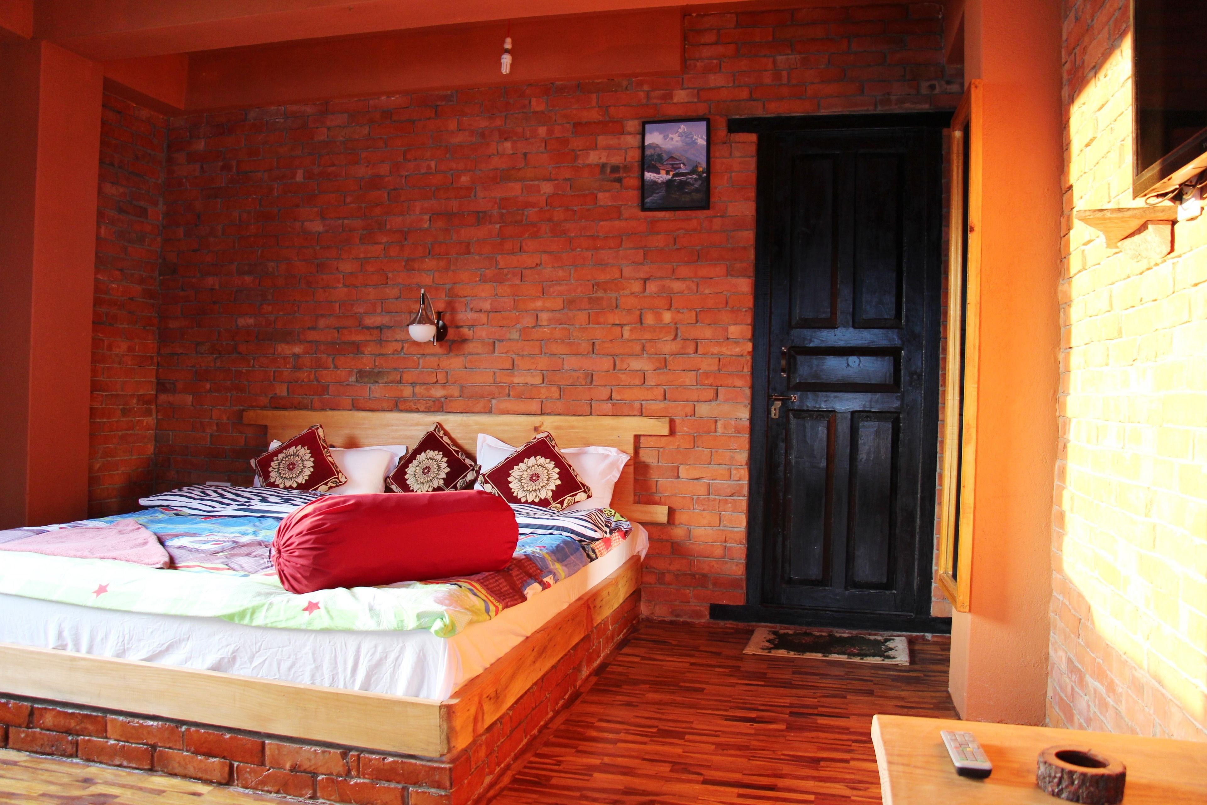 A room with a brick wall, a wooden bed with pillows and a blanket, a wooden table with a remote control, and a black door.