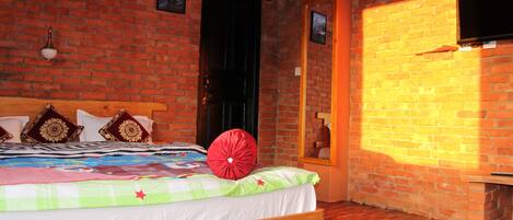 Una habitación con pared de ladrillo, una cama con cabecera de madera, un televisor montado en la pared y una ventana por donde entra la luz del sol proyectando sombras.
