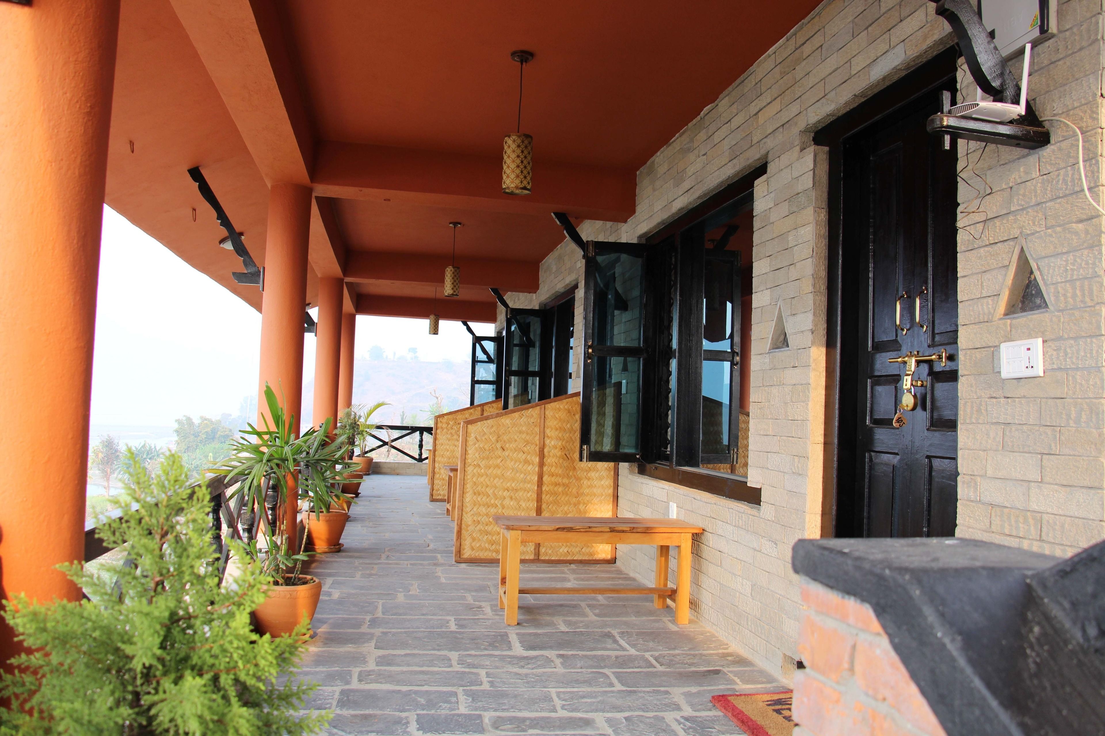 A covered outdoor area with a stone floor, wooden benches, potted plants, and a brick wall with a black door.