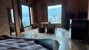 A hotel room with a bed, a dining table with chairs, a refrigerator, and large windows with a view of the sea and mountains. - Nar-Bis Hotel (Pokhara)