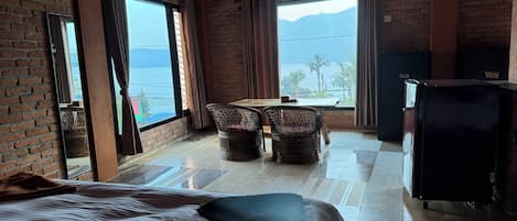 A hotel room with a bed, a dining table with chairs, a refrigerator, and large windows with a view of the sea and mountains.