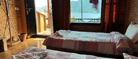 A room with two beds, a wicker chair, a basket, and a view of a mountain and lake through a window.