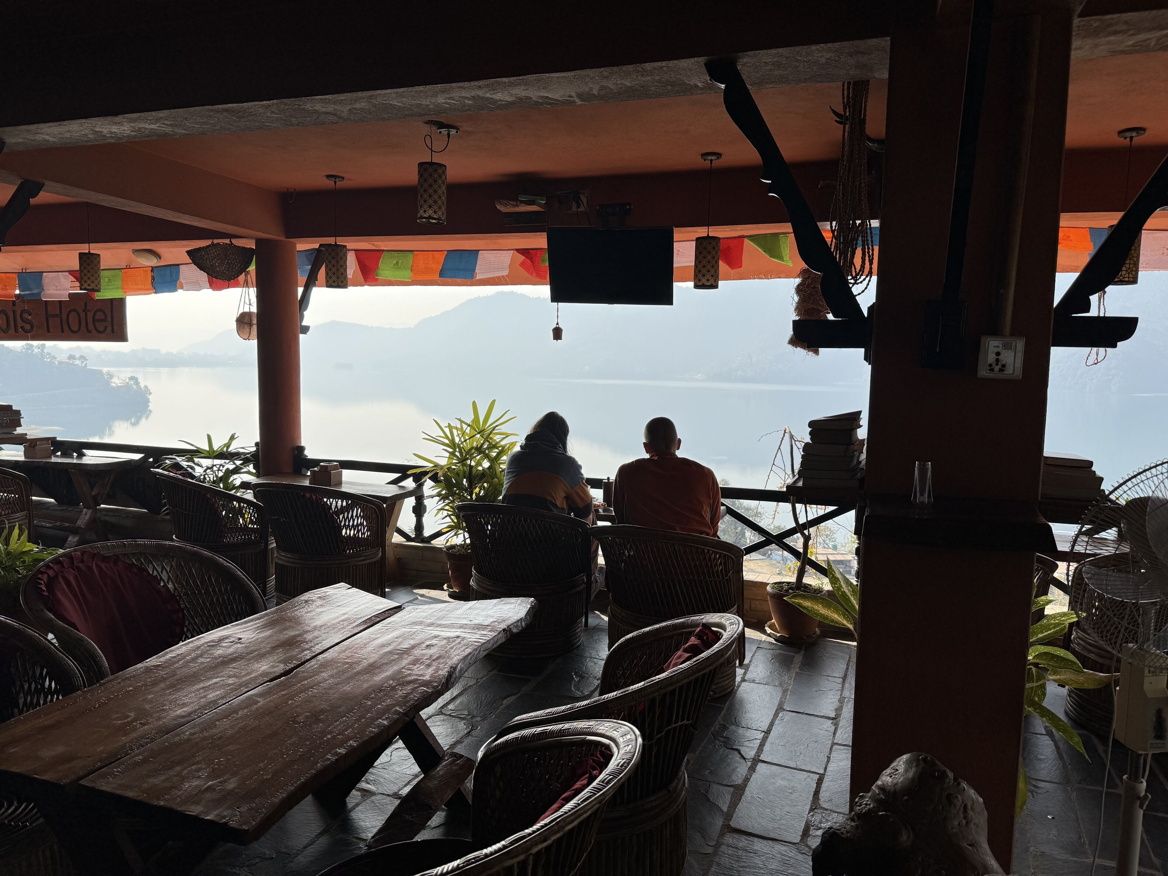 A restaurant with outdoor seating overlooking a lake, featuring wooden tables and chairs, and a view of mountains in the distance.