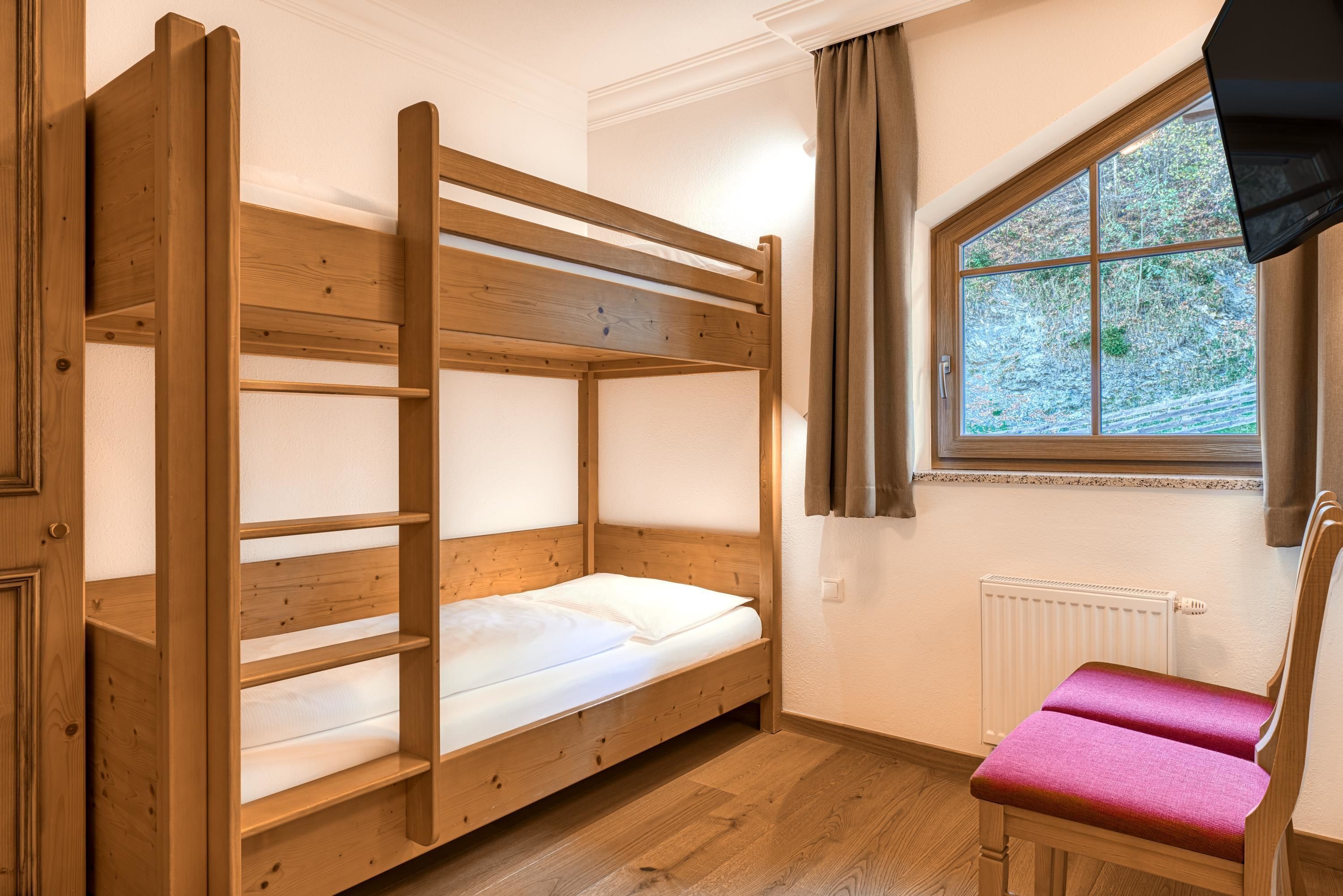 A bunk bed with a wooden frame and white bedding, a window with a view of trees, and a radiator on the wall.
