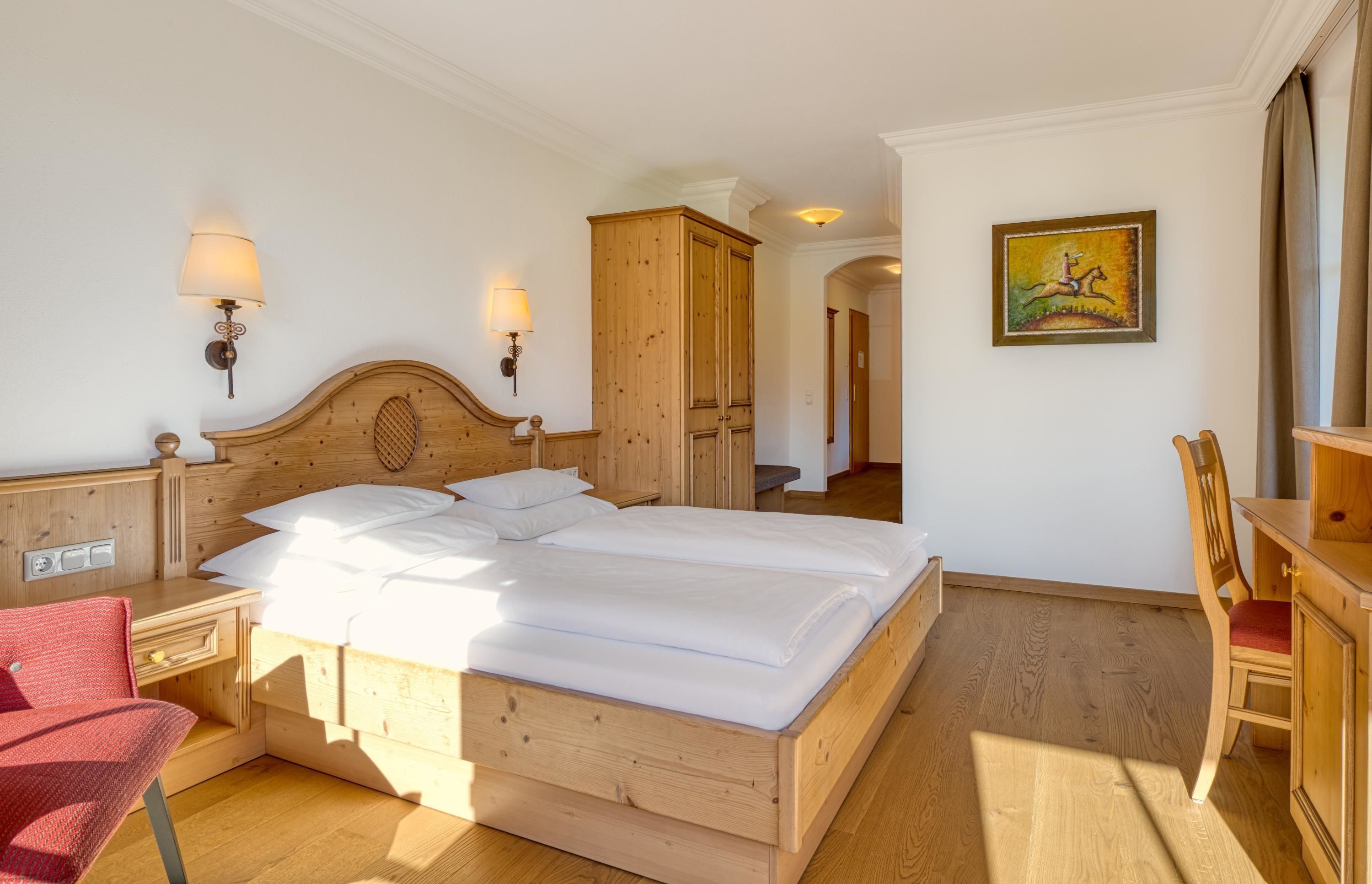 Ein hölzernes Bett mit weißen Leinen, ein Nachttisch aus Holz mit einer Lampe, ein roter Sessel und ein Holztisch mit einem Stuhl.