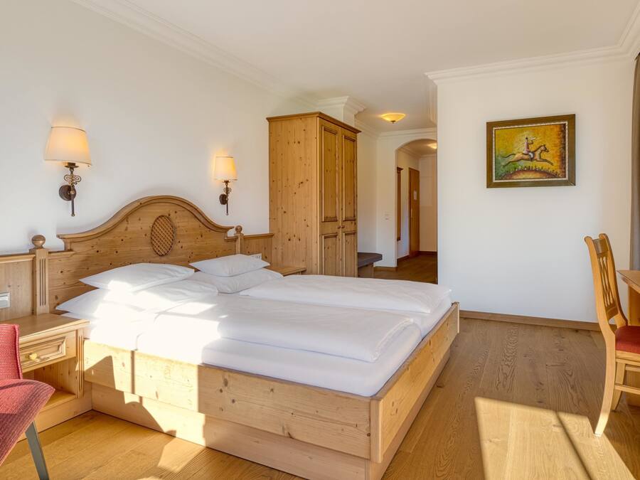 A wooden bed with white linens, a wooden nightstand with a lamp, a red chair, and a wooden desk with a chair.