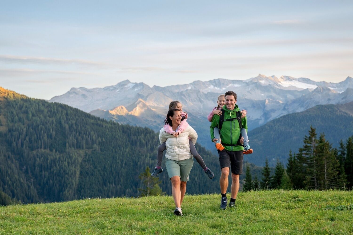 Una familia caminando por la cima de una montaña cubierta de hierba, con montañas nevadas al fondo.