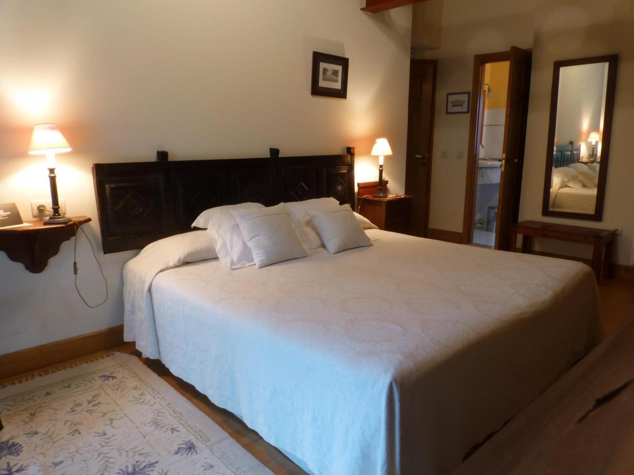 Una cama bien hecha con sábanas y almohadas blancas, dos lámparas de noche, un cuadro en la pared y un piso de madera.
