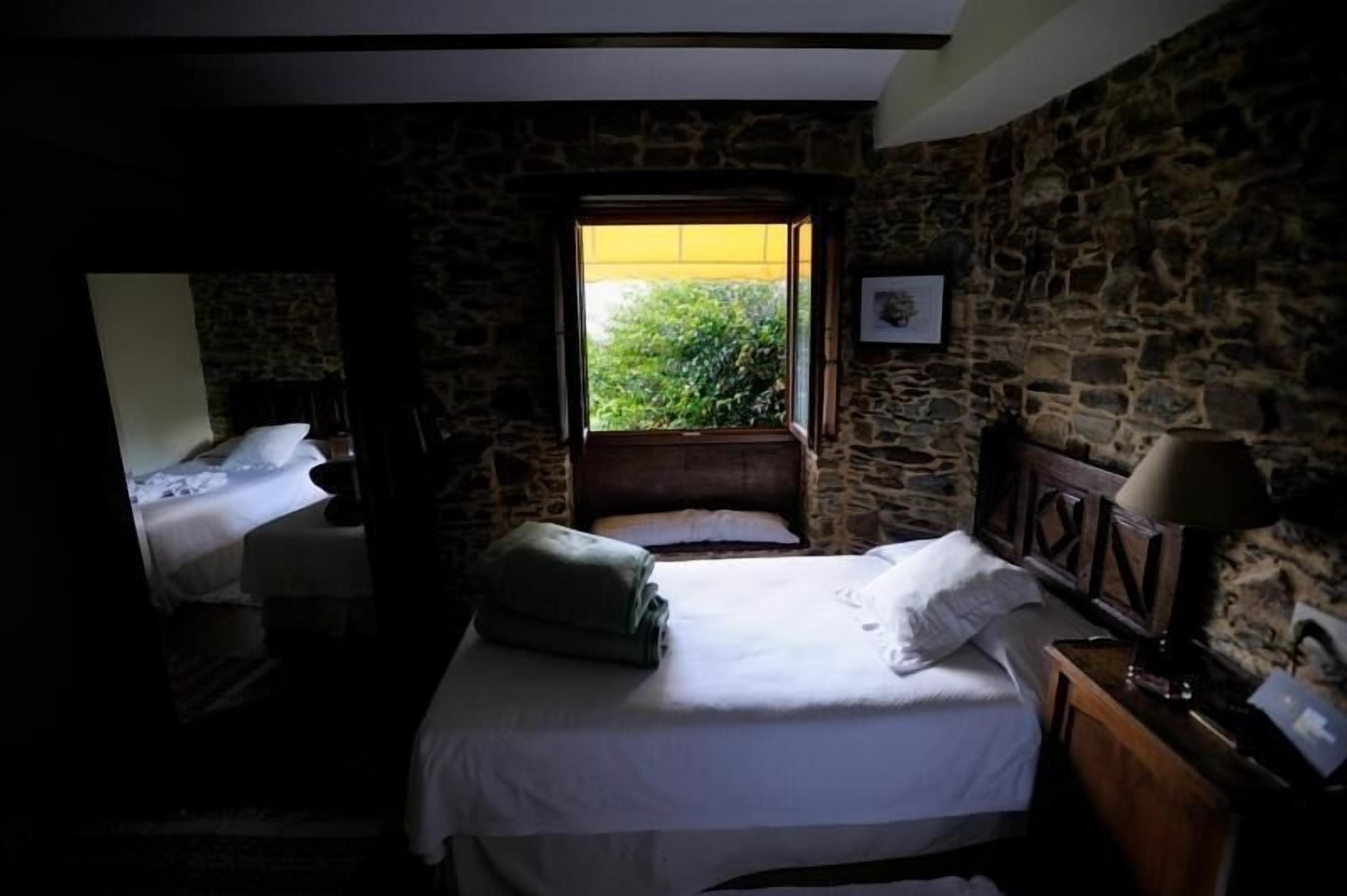 Un dormitorio con pared de piedra, una ventana con vistas a la vegetación, una cama con ropa blanca y una mesita de noche con lámpara.
