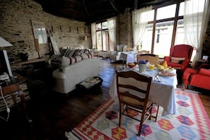 Une pièce rustique aux murs de pierre, avec un canapé, une table à manger recouverte d’un tapis et un fauteuil rouge.