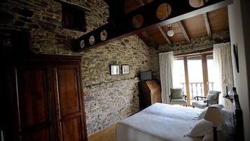 A rustic room with a stone wall, wooden beams, a bed, a wardrobe, and a balcony with chairs.