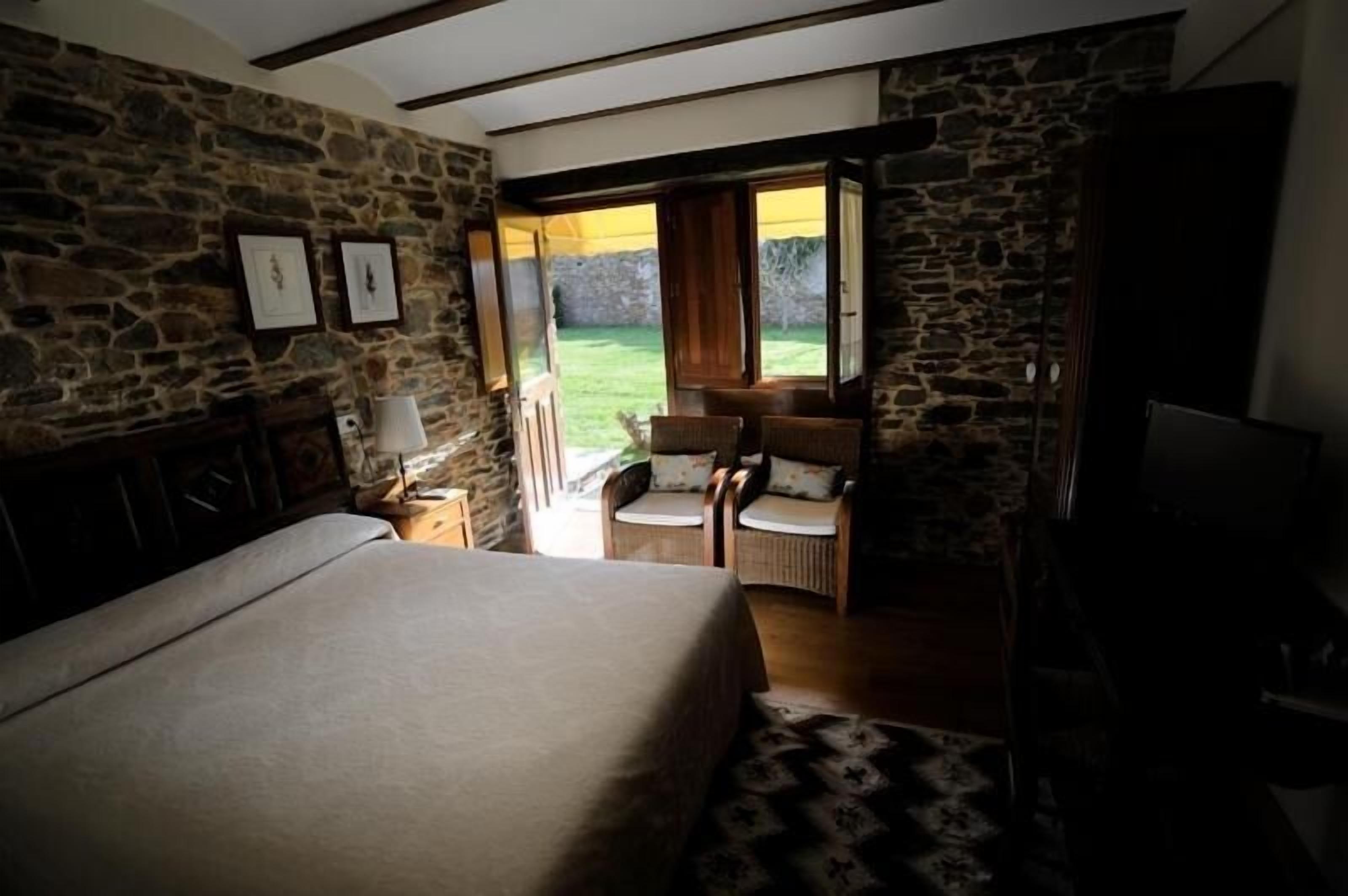 A bedroom with a large bed, a wooden chair, a small table with a lamp, and a stone wall.