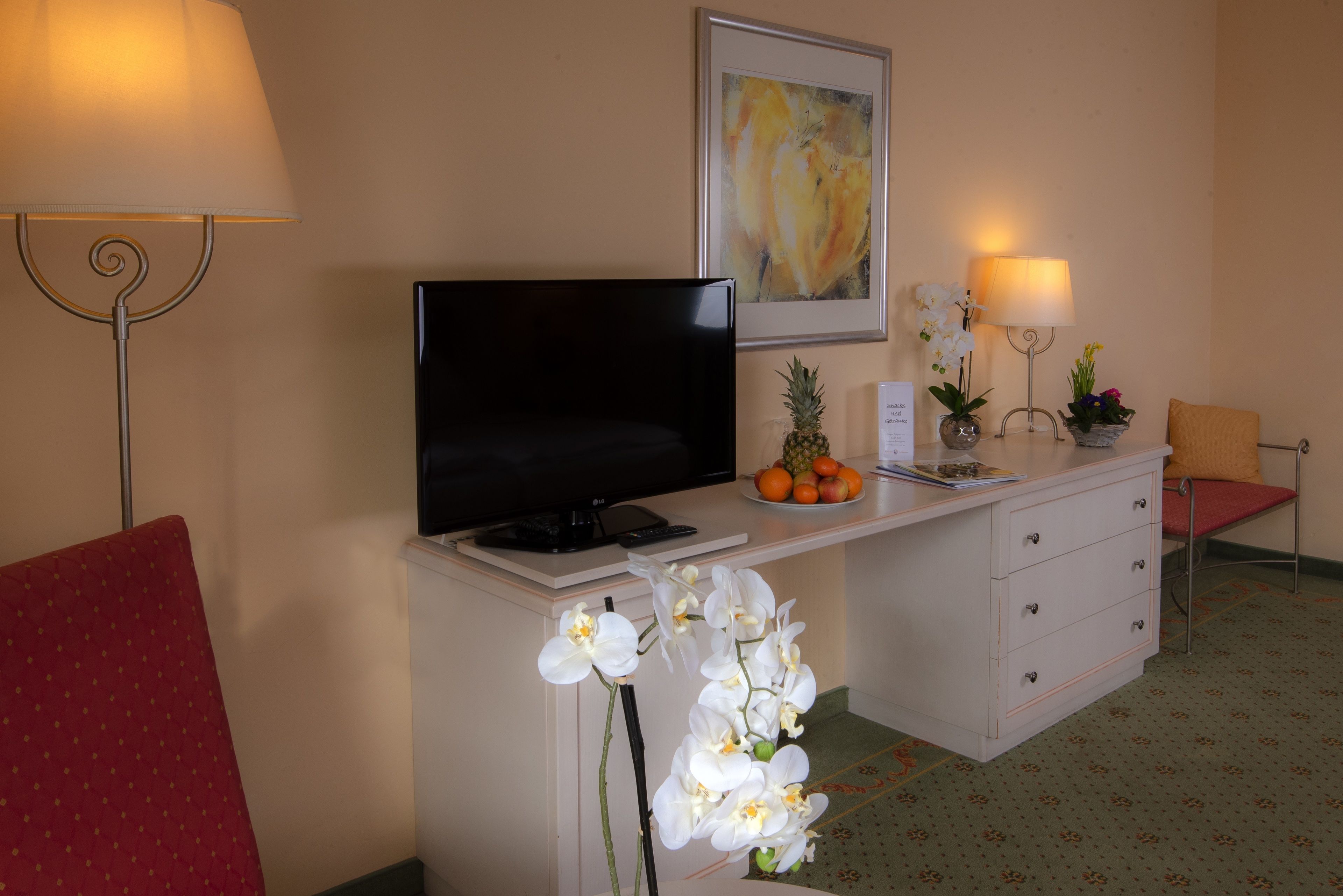 A hotel room with a TV, a desk with a laptop, a plant, and a framed picture on the wall.