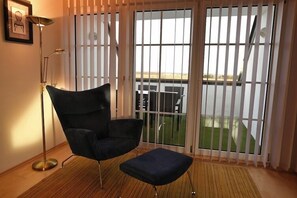A room with a dark armchair and ottoman, a floor lamp, and a view through a large window with blinds. - Thermenhotel 4 Jahreszeiten (Lutzmannsburg)