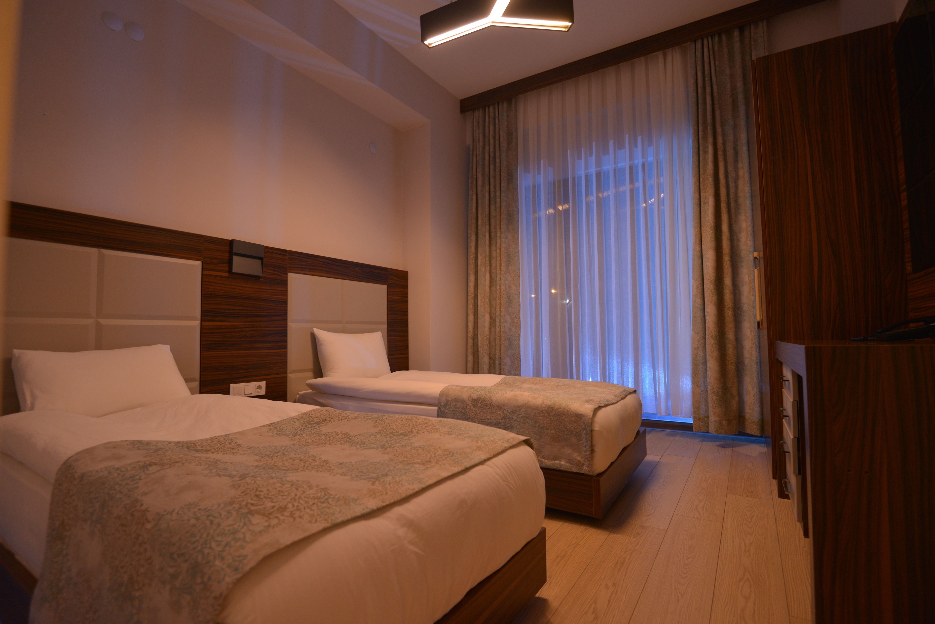 Habitación de hotel con dos camas, cabecero de madera, una ventana grande con cortinas y suelo de madera.