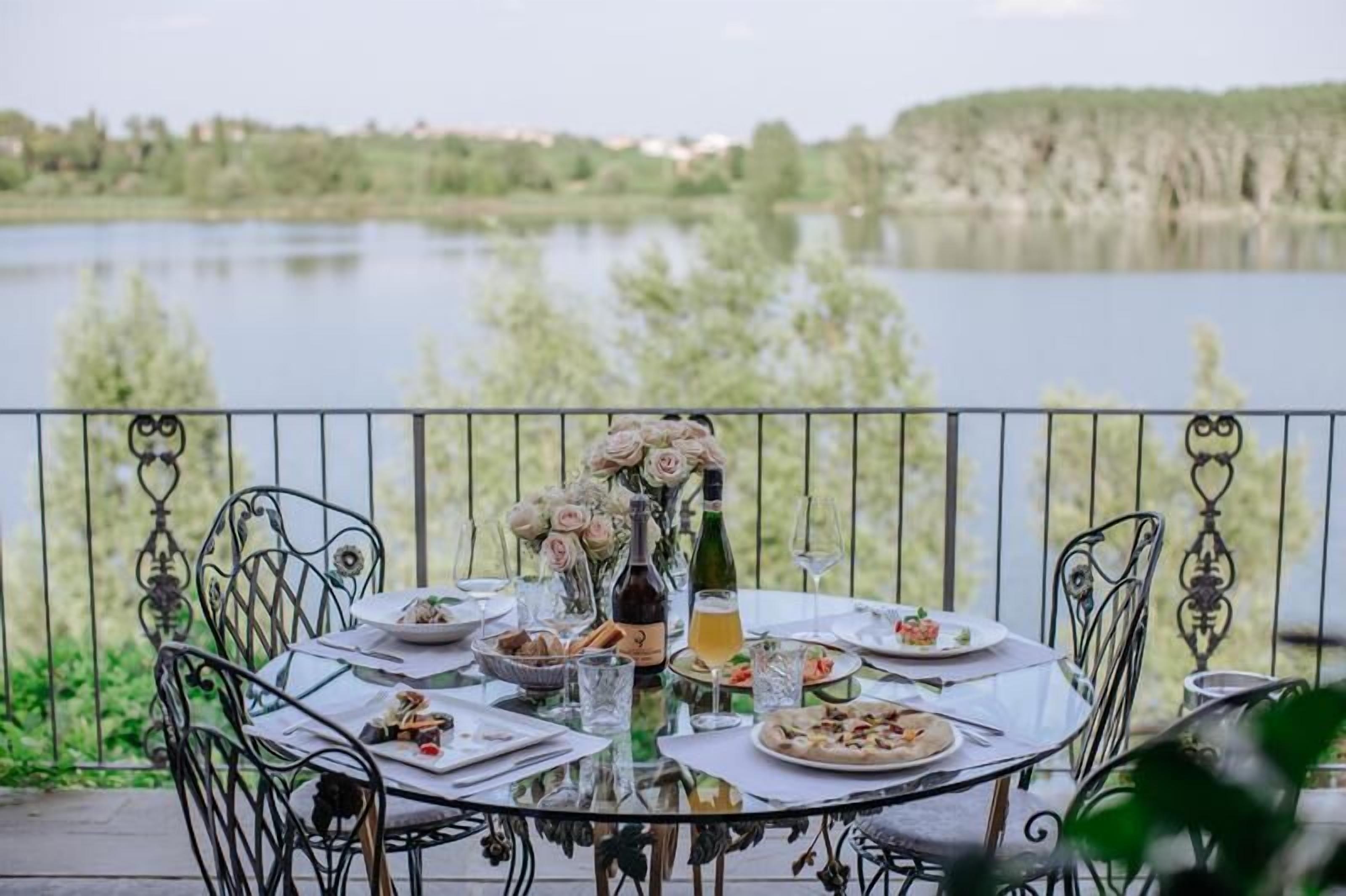 A round dining table set outdoors with a view of a lake, surrounded by metal chairs.